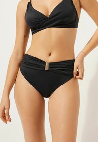 Svart bikiniset med en topp i inslagningstil och högmidjade bottnar. Har en samlad textur och metallspänne vid midjan.