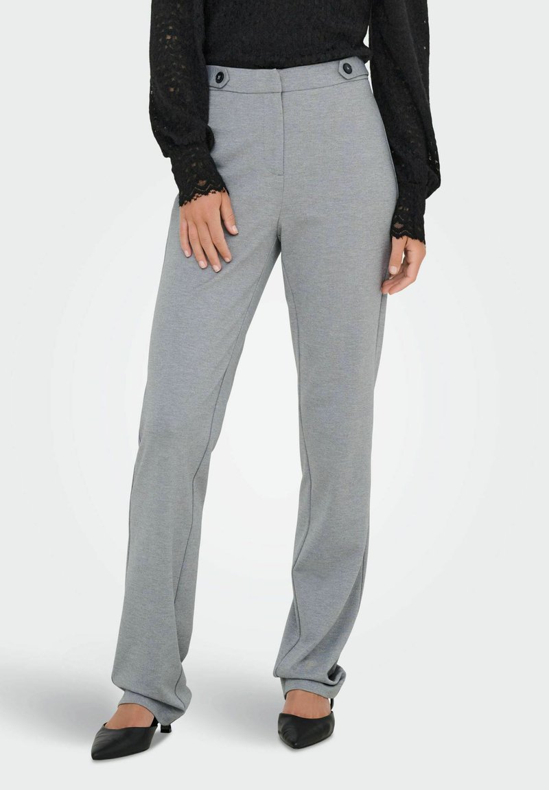 Femme portant un pantalon tailleur gris clair avec des détails de boutons et des chaussures noires à bout pointu, associé à un haut en dentelle noir à manches longues.
