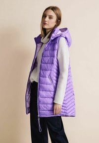 Lila puffarväst med huva, med horisontell quiltad design och framfickor med dragkedja. Bärs över en ljusgrå tröja och mörka byxor.