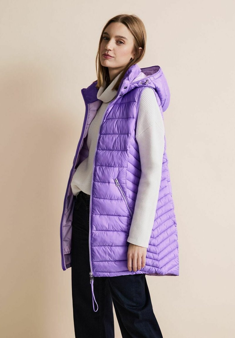 Lila puffarväst med huva, med horisontell quiltad design och framfickor med dragkedja. Bärs över en ljusgrå tröja och mörka byxor.