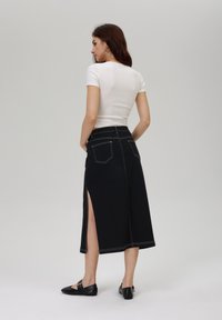Jupe en denim noire avec une fente haute sur un côté et des coutures blanches contrastantes, associée à un t-shirt blanc ajusté et des ballerines noires.