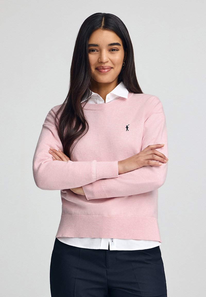 Maglione a maglia rosa chiaro con maniche lunghe, collo rotondo e un piccolo logo nero. Indossato sopra una camicia bianca con colletto, abbinato a pantaloni scuri.