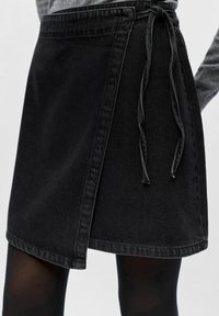 Jupe portefeuille en denim noir avec lien sur le côté, portée avec des collants noirs et un haut à manches longues gris.