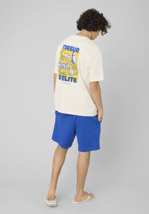 Homme debout de dos, portant un t-shirt crème avec un motif pétanque coloré et un short bleu, mains dans les poches, sur fond gris.