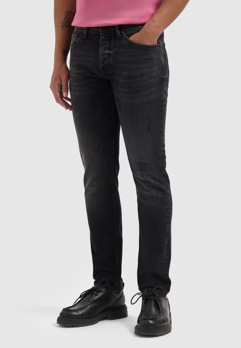 Zwarte spijkerbroek met een slim fit, voorzien van subtiele vervaging, een versleten patch op het been en een klassieke vijf-pocketontwerp, gecombineerd met zwarte schoenen.