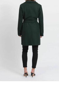 Dunkelgrüner Wollmantel mit tailliertem Gürtel, langen Ärmeln und Reverskragen, kombiniert mit schwarzen, schmal geschnittenen Hosen und hochhackigen Schuhen.