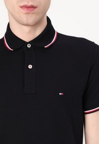 Polo negro con una tela texturada, que presenta un cuello acanalado con rayas rojas y blancas, y una pequeña insignia de logo en el pecho.