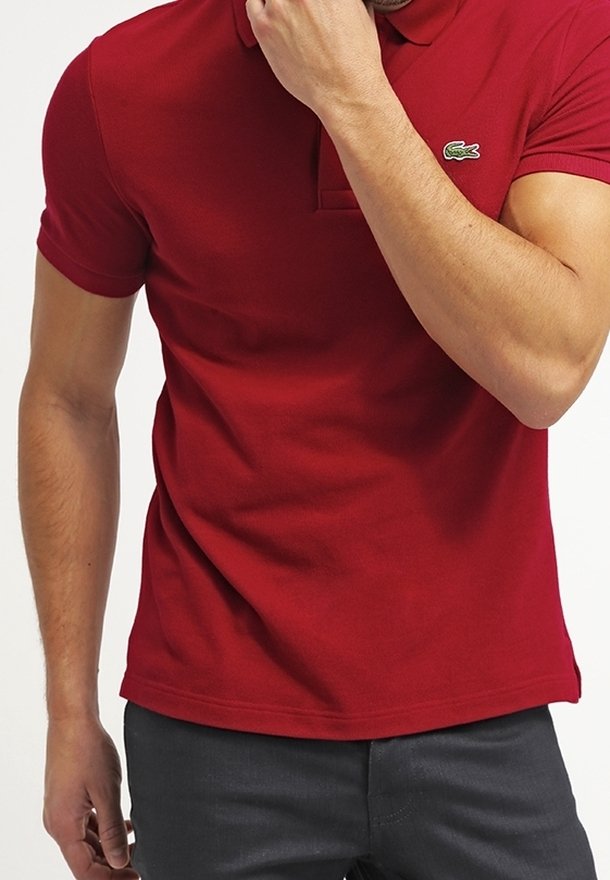 Homme portant un polo rouge ajusté avec un petit logo crocodile vert sur la poitrine, debout avec une main près du visage.