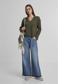 Sweter z dzianiny w oliwkowej zieleni z dekoltem w serek i długimi rękawami, w zestawie z szerokimi niebieskimi dżinsami. Uzupełniony o wzorzysty szalik.