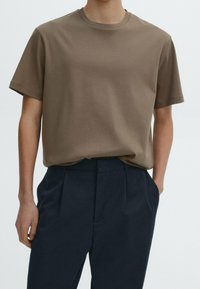 Man wearing a plain brown short-sleeve t-shirt tucked into high-waisted black pleated trousers, hands in pockets.