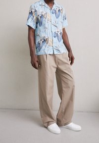 Camicetta a maniche corte blu con stampa floreale e foglie di palma, abbinata a pantaloni beige morbidi e scarpe da ginnastica bianche. Tessuto morbido con vestibilità rilassata.