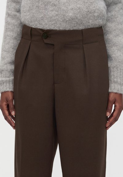 Personne portant un pantalon marron taille haute avec plis à l'avant et un seul bouton, associé à un pull en maille gris clair.