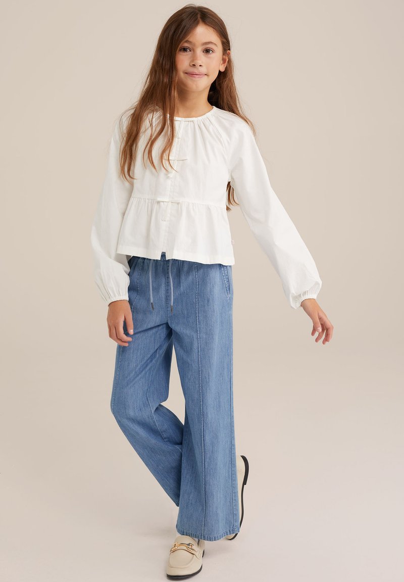 Top peplum alb din bumbac, cu mâneci lungi; pantaloni denim blue cu croială largă; stilizat cu încălțăminte ușoară, cu un accent decorativ.