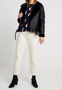 Black leather motorcycle jacket with shearling collar, paired with a navy patterned shirt, cream cropped jeans, and black heeled ankle boots.