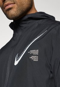 Μαύρο αθλητικό μπουφάν με χαρακτηριστικό λευκό λογότυπο Nike και λεπτομέρειες κειμένου, με φερμουάρ μπροστά και κουκούλα. Λείος υφασμάτινος συνδυασ μός.