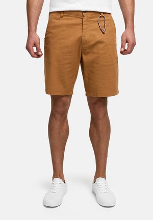Mann trägt braune knielange Shorts mit einem geflochtenen Schlüsselanhänger, weiße Sneaker und ein weißes Hemd, steht vor einem weißen Hintergrund.