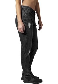 Jeans de mezclilla negra con un corte relajado, que presentan parches desgastados y bordes deshilachados. Combinados con zapatillas deportivas negras.