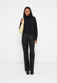 Schwarzer Rollkragenpullover kombiniert mit hoch taillierten schwarzen Hosen. Hält eine blassgelbe Handtasche. Glatter Stoff mit taillierter Passform.