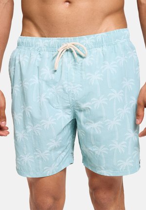 Hombre con pantalones cortos de baño azul claro con patrones de palmeras blancas y un cordón blanco atado en la cintura.
