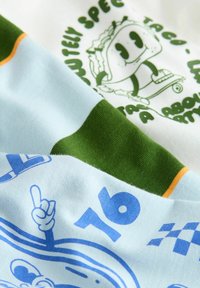Közelkép egy zöld és világoskék csíkos anyagról, kék 16-os számmal, egy kézzel, amely kő löketet mutat, valamint egy zöld, rajzfilm tacoval, amely gördeszkán áll.