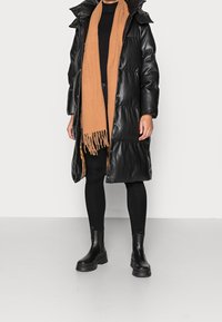 Černý péřový kabát s lesklou texturou, béžový šátek s třásněmi, přiléhavý černý top, černé legíny a černé kotníkové boty s tlustými podrážkami.