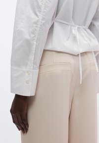 Balta krekla ar garām piedurknēm un sasienamu jostasvietu, ar apaļiem manšetiem un pogu detaļām. Kombinēts ar bēšām, piegrieztām biksēm ar aizmugurējām kabatām.