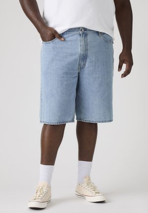Man draagt lichtblauwe denimshorts, witte sokken, beige sneakers en een wit t-shirt met één hand in de zak.