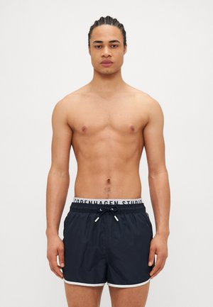 Marineblaue Badeshorts mit weißem Besatz und Kordelzug, ausgestattet mit einem elastischen Bund, der mit "COPENHAGEN STUDIO" beschriftet ist. Glattes Material, sportlicher Schnitt.