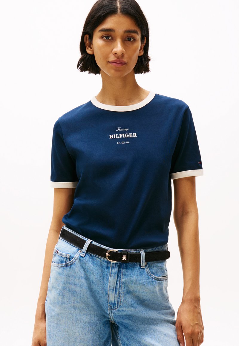 T-shirt à manches courtes navy avec col et poignets côtelés blancs. Présente un logo textuel sur le devant et est associé à un jean bleu clair et une ceinture noire.