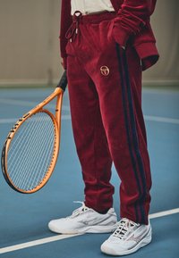 Bordo velūra sporta bikses ar zilu sānu svītru un zelta logo; kopā ar baltām sporta apaviem un turot oranžu tenisa raketi.