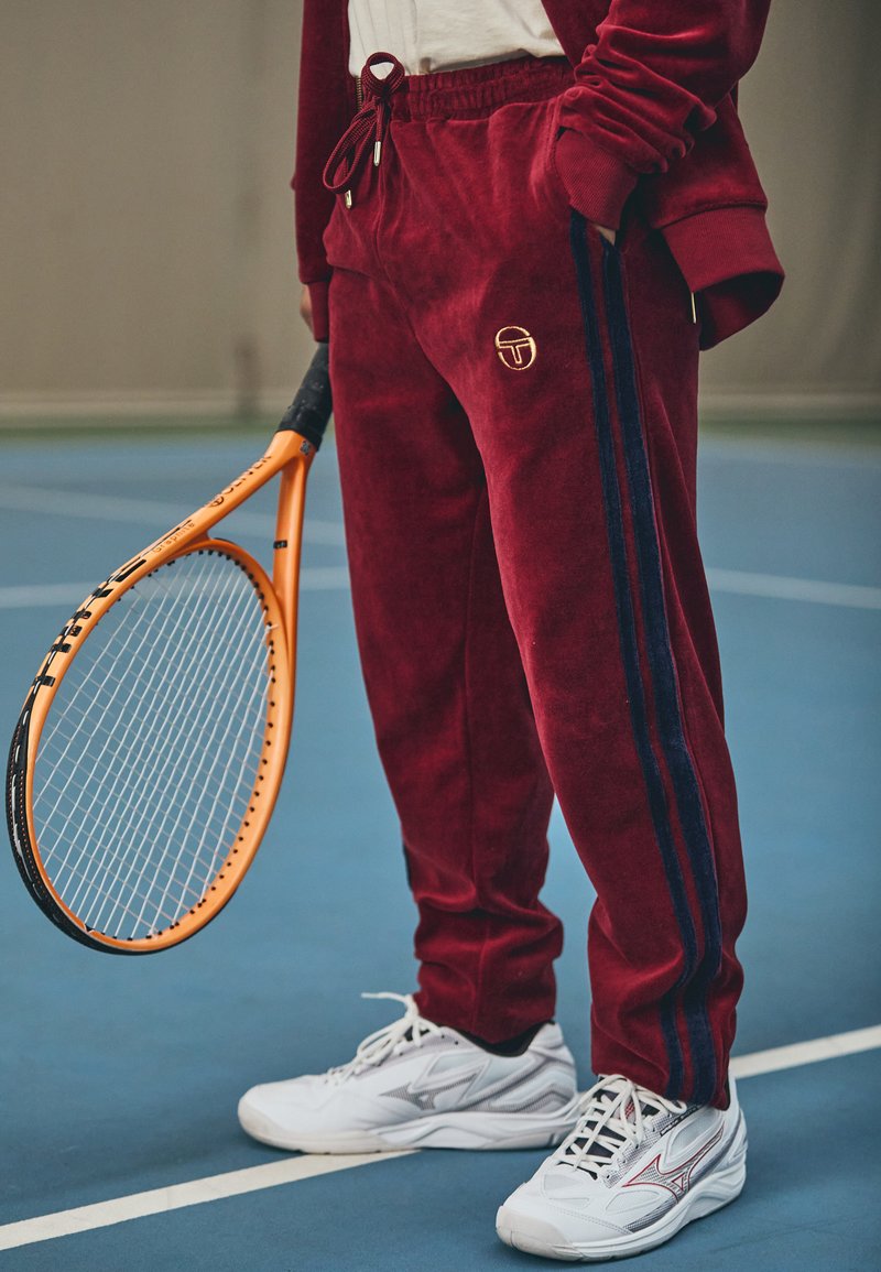 Bordo velūra sporta bikses ar zilu sānu svītru un zelta logo; kopā ar baltām sporta apaviem un turot oranžu tenisa raketi.