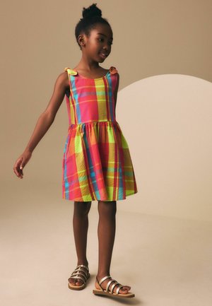 Young girl in a colorful plaid dress and gold sandals, standing and looking to the side against a beige background.