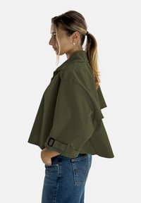 Olivgrön croppad jacka med krage, knappstängning och skärpade manschetter. Slät textur och flödande tyg med lager på lager-detalj på ryggen.