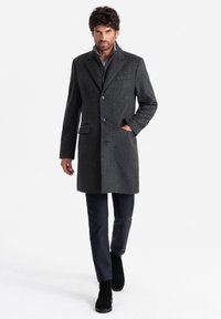 Abrigo gris de espiga con tres botones, dos bolsillos laterales y solapas de muesca, combinado con pantalones oscuros y botas negras.