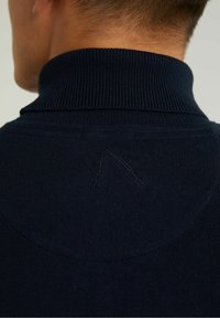 Marinblå stickad tröja med hög krage, med en liten sömnadsdetalj vid nacken och en slät textur.