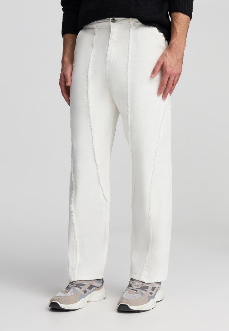 Pantaloni in denim bianco con vestibilità dritta, caratterizzati da dettagli di cucitura in evidenza e orlo sfrangiato. Abbinati a scarpe sportive grigie e beige.