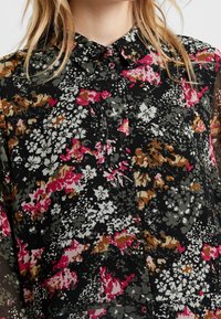 Chemise noire à boutons avec un motif floral coloré en rose, gris et blanc. Fabriquée en tissu léger avec une finition texturée.