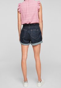 Haut rose clair avec manches volantes, associé à un short en denim foncé avec ourlets retroussés et taille élastique. Le mannequin porte des chaussures blanches.