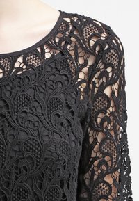 Top in pizzo nero caratterizzato da intricati motivi floreali e swirl, con maniche lunghe e scollatura rotonda, che offre un design decorativo e testurizzato.