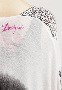 Top ligero de punto blanco con un patrón abstracto negro en el hombro y un logo "Desigual" en rosa, con textura suave y corte relajado.