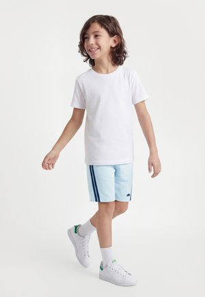 Usmievajúce sa dieťa v bielom tričku, svetlomodrých šortkách s tmavomodrými pruhmi, bielych ponožkách a bielych teniskách so zelenými detailmi, kráčajúce dopredu.