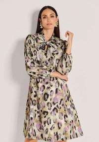 Frau, die ein langärmliges, kniebedeckendes Kleid mit Leopardenmuster und einem großen Schleifenkragen sowie auffälligen goldenen Ohrringen trägt.