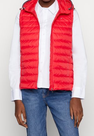 Person iført en skarpt rød quiltet ærmeløs hættetrøje over en hvid skjorte med knapper og blå jeans, hænder afslappet langs siden.