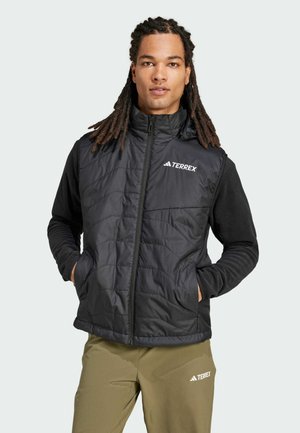 Gilet noir isolé avec un design matelassé, un col haut et un logo sur la poitrine, associé à un haut noir à manches longues. Coupe ajustée.