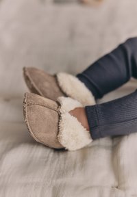 Stópki niemowlęcia w miękkich beżowych zamszowych kapciach z białym futerkiem, ubrane w ciemnoniebieskie prążkowane spodnie, spoczywające na kremowym kocu.