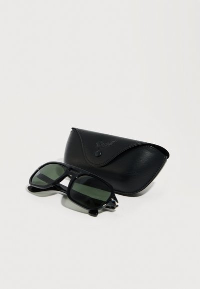 Γυαλιά ηλίου με μαύρο πλαίσιο και σκουρόχρωμους φακούς τοποθετημένα δίπλα σε μια μαύρη δερμάτινη θήκη σε λευκό απλό φόντο.