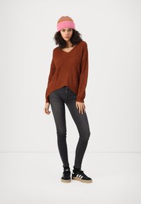Roestkleurige gebreide trui met V-hals, gecombineerd met zwarte skinny jeans en zwarte sneakers met witte strepen. Roze en bruine gestreepte beanie.