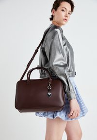 Brown leather handbag with a structured shape, featuring a top handle and an adjustable shoulder strap. Includes a heart charm detail.