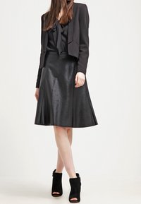 Blazer nero corto su una blusa nera con scollatura annodata, abbinato a una gonna nera in pelle svasata e stivali alla caviglia neri con punta aperta.