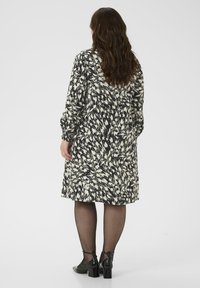 Vrouw met lang golvend haar, gekleed in een knielange zwart-witte jurk met een patroon, zwarte doorschijnende panty en zwarte hakken, staand met haar rug naar de kijker gekeerd.